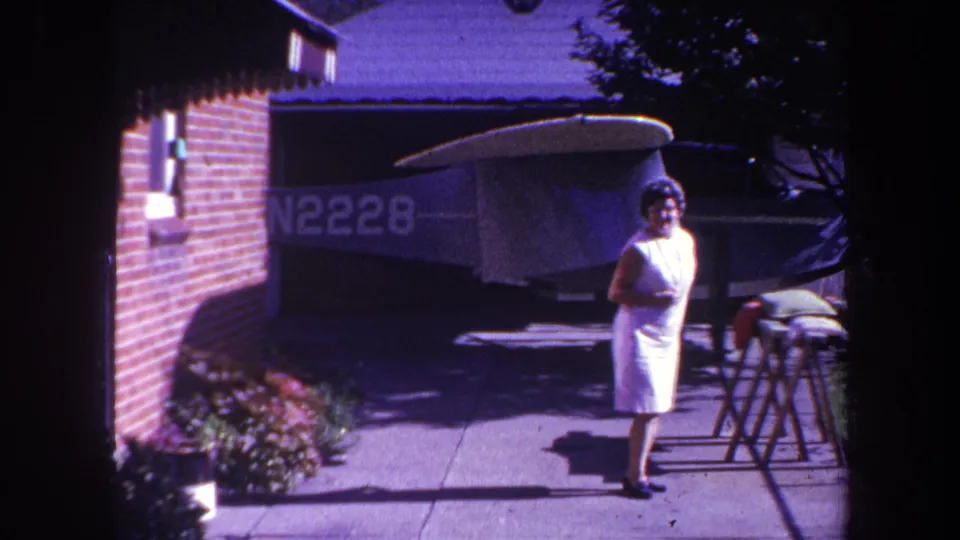 1969 Yucaipa frame with a woman in a white dress near a shaded driveway, brick wall, shrubs, and a covered light aircraft marked N2228.