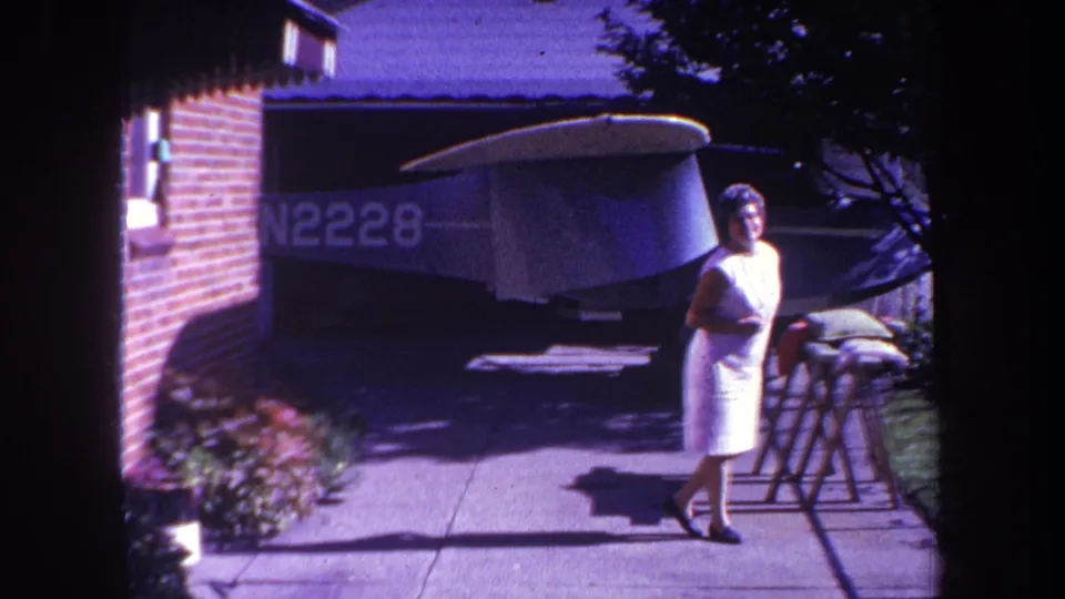 1969 Yucaipa home-movie frame showing the woman on the sidewalk, with the aircraft tail and N2228 marking partly visible behind her.