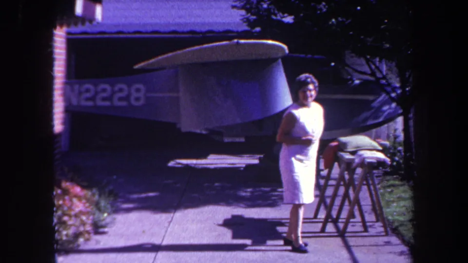 1969 Yucaipa frame showing the woman near wooden supports, sidewalk, lawn, brick house, and the shaded aircraft parked behind her.