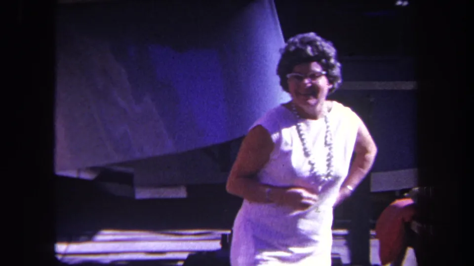 A smiling woman in a white dress and glasses gestures toward the camera beside a large aircraft form in 1969 California home-movie footage.