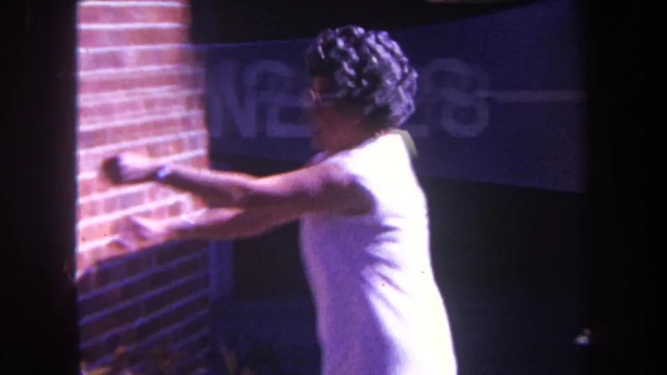 The woman turns sideways and reaches toward a brick exterior wall, with a dark doorway or window behind her in the 1969 home-movie clip.