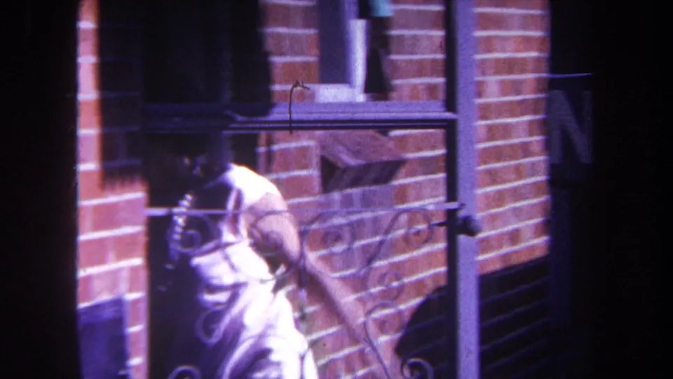 A woman in a white dress moves beside a brick building with a window and decorative metal railing in 1969 Yucaipa-area footage.