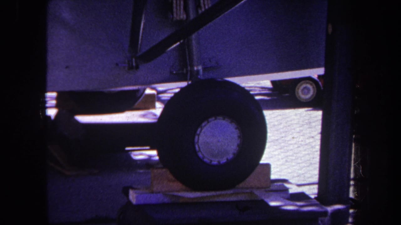 1969 8mm frame from Volmer Yucaipa showing a large mobile home wheel, wood blocking, undercarriage shadows, and paved ground.