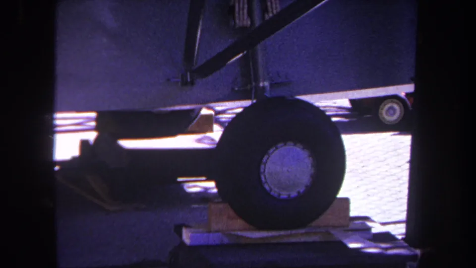 1969 8mm close view of a mobile home wheel supported near wooden blocks, with railing and paved ground visible around the undercarriage.