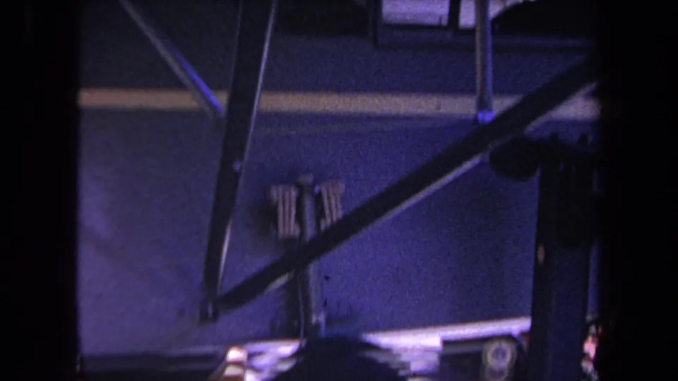 Later 1969 frame shows the mobile home underside from a low angle, with diagonal railing, partial wheel area, wood block, and bright pavement.