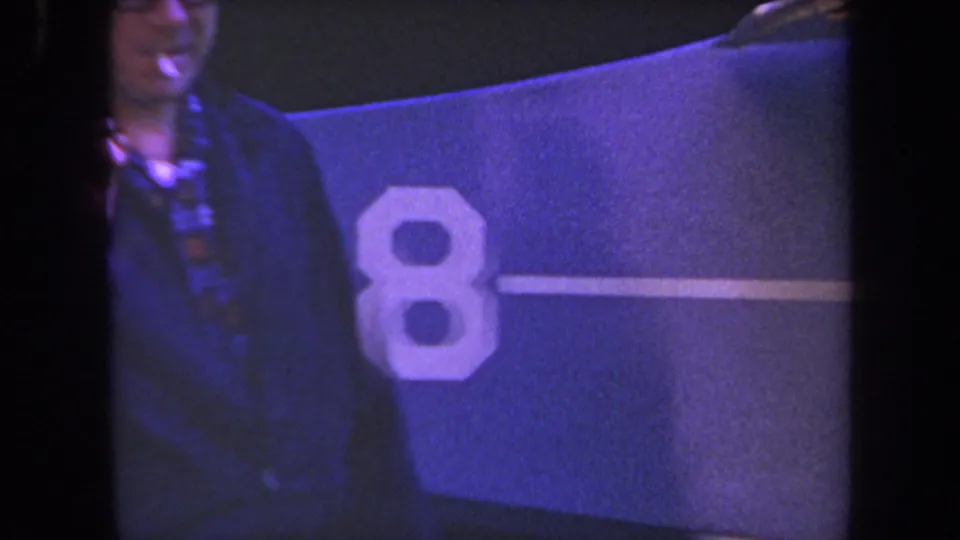 Adult man with glasses and cigarette stands beside a blue aircraft marked with a large number 8 in the 1969 Yucaipa-area footage.