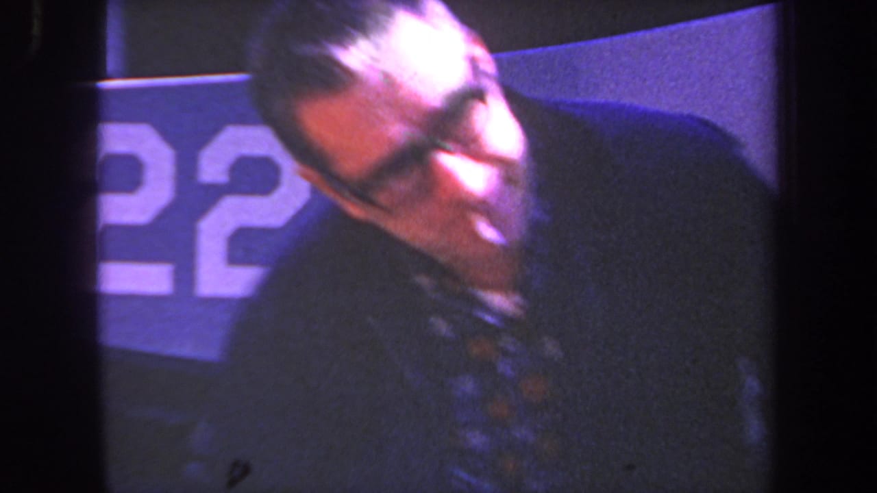 Man with glasses leaning beside a blue surface with partial white numbering.