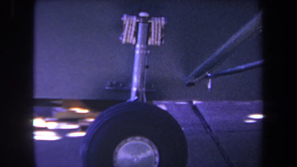 1969 home-movie frame with the aircraft wheel low in frame, a vertical strut above it, and a diagonal support near the fuselage.