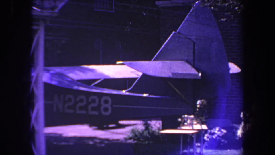 1969 Yucaipa frame showing the tail of a small aircraft, partial registration N2228, brick wall, and nearby foliage.