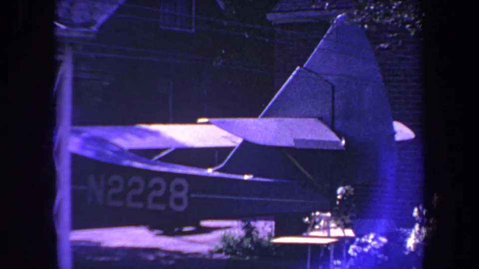 Late-1960s 8mm frame of a small private airplane's tail and rear fuselage, marked N2228, in a residential nighttime setting.