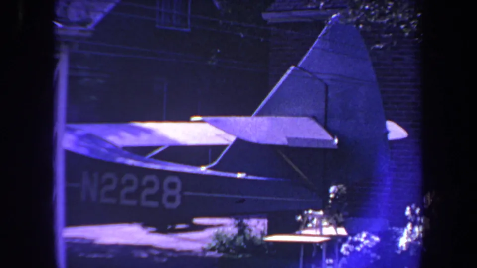 1969 home-movie frame showing a small airplane's rear fuselage and tail near a brick house, with N2228 faintly visible in low light.