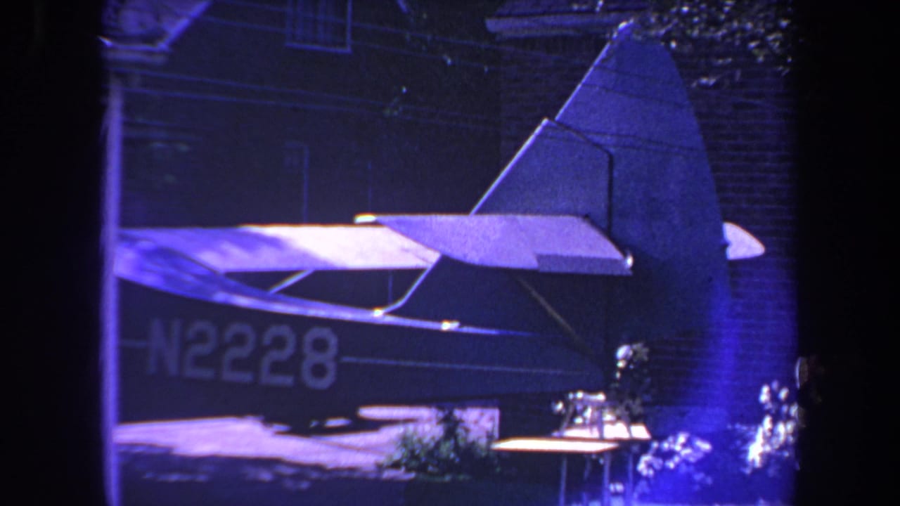 1969 Yucaipa home-movie frame showing the rear of a small airplane with registration N2228 near a brick residence under blue-purple lighting.
