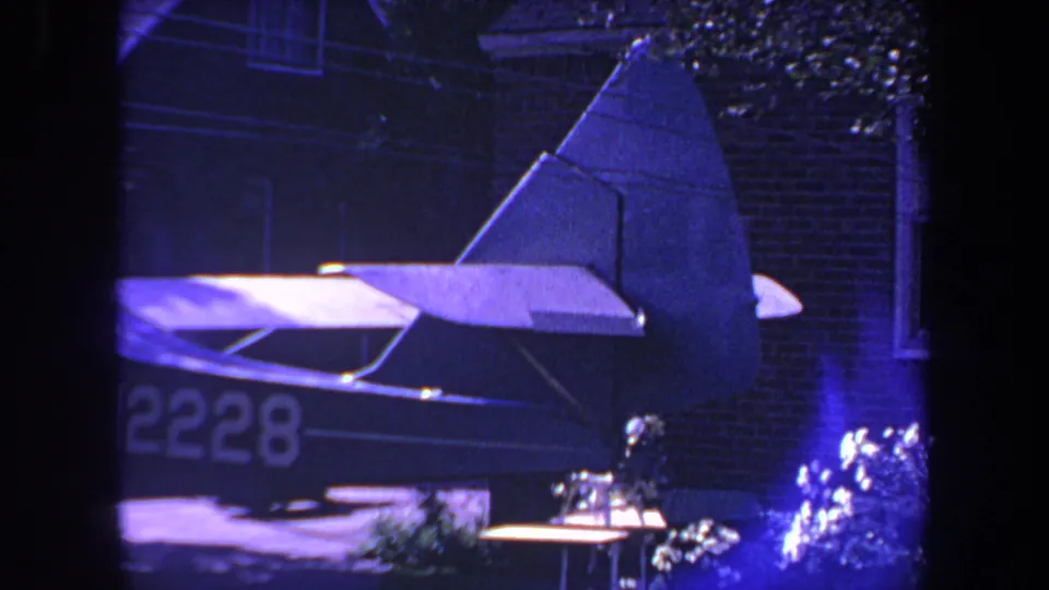 1969 Yucaipa frame showing the rear of a small airplane with partial N2228 marking near a brick house, plants, and patio furniture at night.