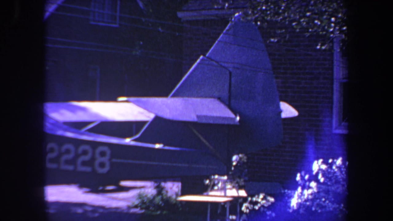 1969 Yucaipa home-movie frame showing a small aircraft tail and wing near a brick house exterior, tinted blue-purple by the film scan.
