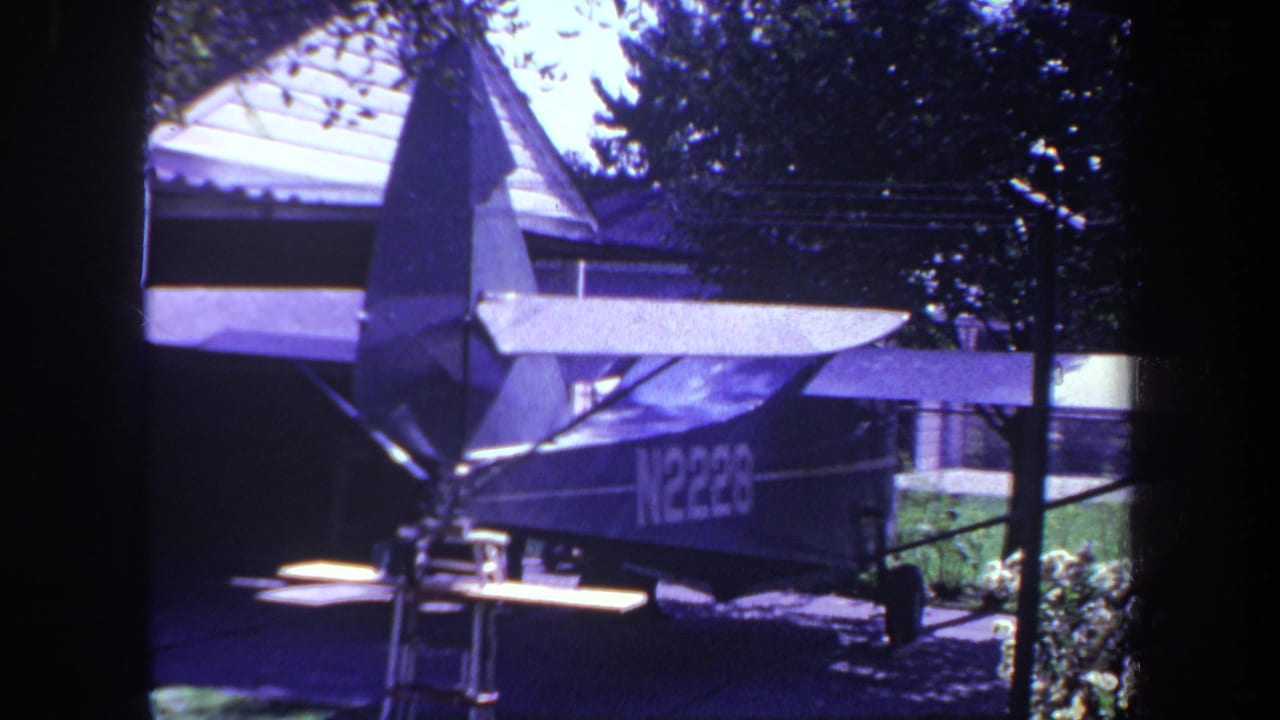 Small high-wing airplane parked in a driveway beside a house and trees.