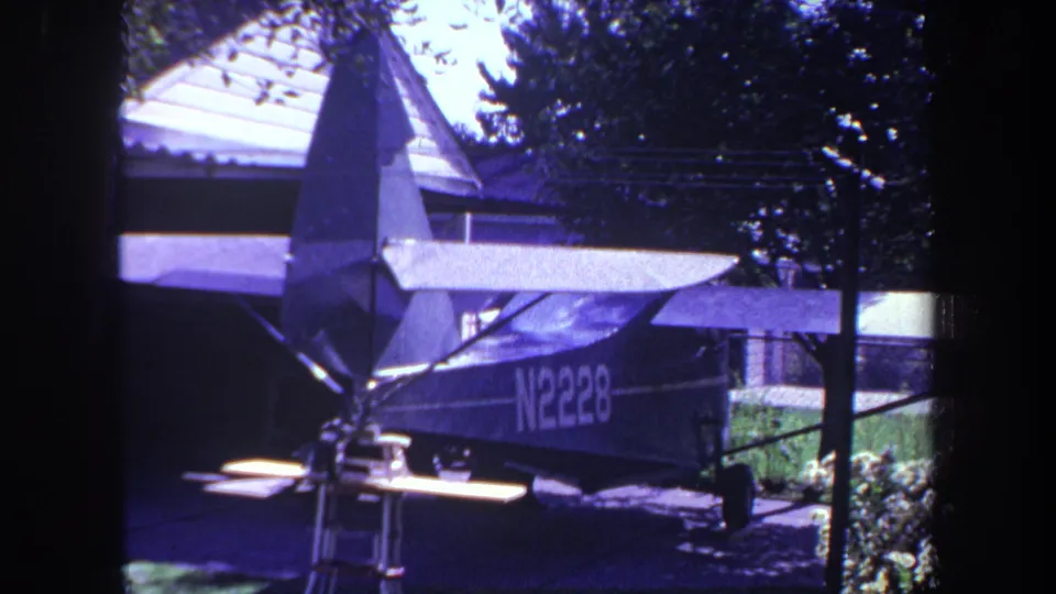 1969 Yucaipa home-movie view of airplane N2228 in a shaded residential driveway with trees, roofline, and planting nearby.