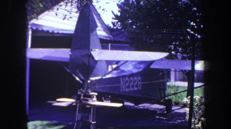1969 Yucaipa frame showing the parked high-wing airplane in a residential driveway, with N2228 marking, trees, and house roof visible.