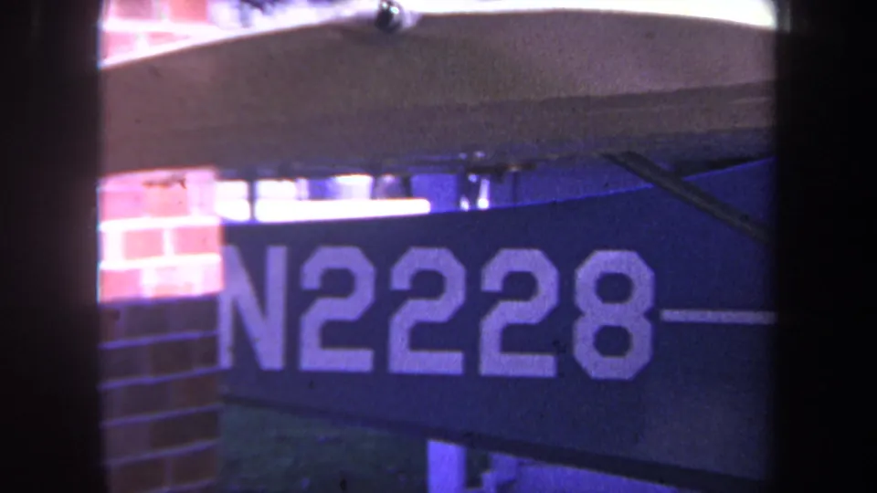 1969 8mm frame from Yucaipa showing the painted aircraft number N2228 near a brick wall and wing structure in bright daylight.