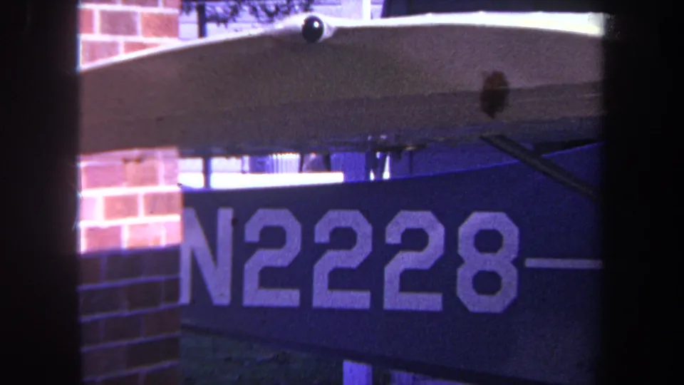 Late-1960s Yucaipa home-movie frame showing the N2228 aircraft marking, a wing edge, support strut, and brick wall background.