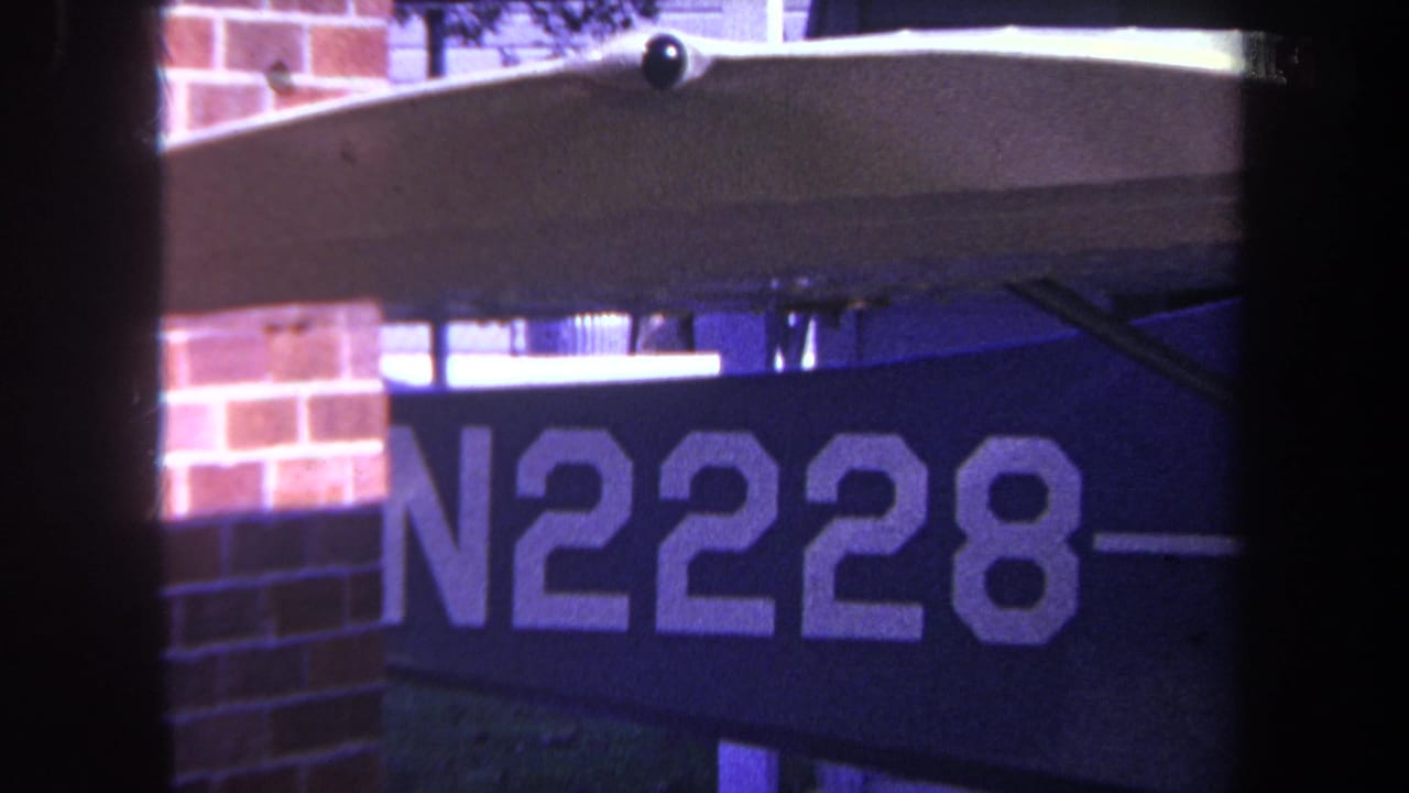 1969 Yucaipa 8mm frame showing a dark blue sign with white N2228 lettering mounted near brickwork, with heavy gate framing.