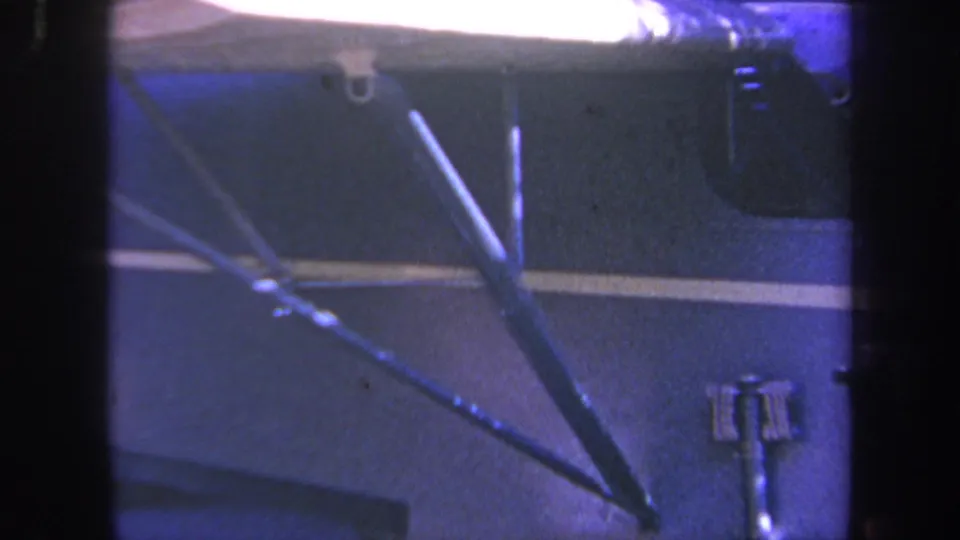 1969 Volmer Yucaipa home-movie frame showing a dark blue vehicle side, white stripe, and metal workshop arms under blue-tinted light.