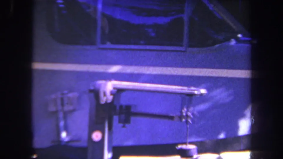 1969 workshop frame showing a dark vehicle body with white stripe and a horizontal mechanical arm or tool in the foreground.