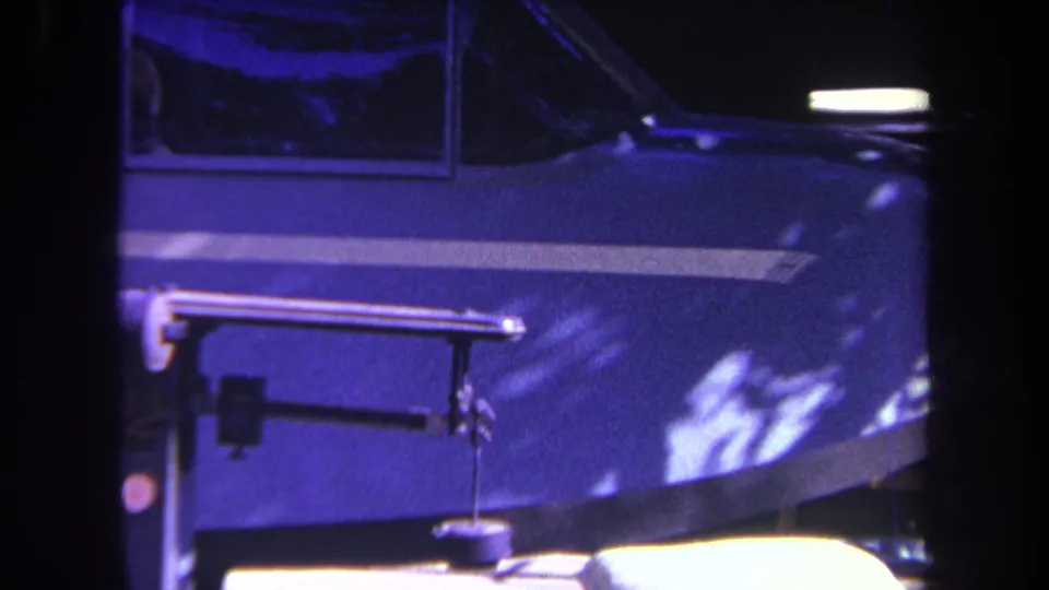 Final blue-tinted 8mm frame with a mechanical arm in front of a dark blue vehicle side and bright light along the lower edge.