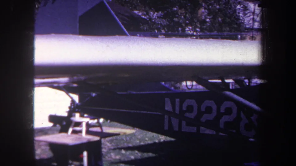 1969 Yucaipa-area 8mm frame showing a parked small aircraft wing and dark tail section with partial N2228 registration near a brick wall.