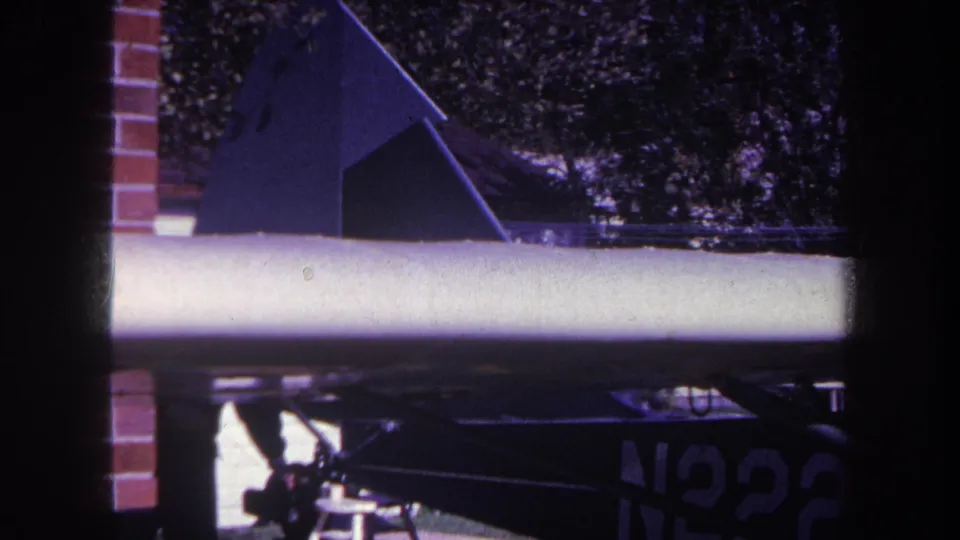 1969 Yucaipa-area home-movie frame showing a small aircraft wing, dark tail section, partial registration, and trees behind it.