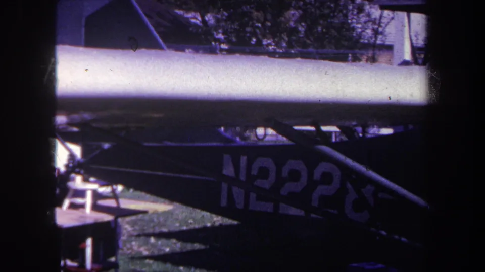 1969 Yucaipa 8mm frame showing the aircraft wing, partial N2228 registration, dark struts, grass, and a small stool-like object near the plane.