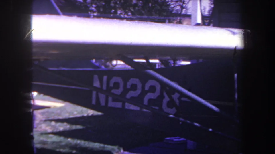 1969 Yucaipa 8mm frame with a pale aircraft wing, partial N2228 registration, dark bodywork, trees, grass, and blue roof forms in the background.