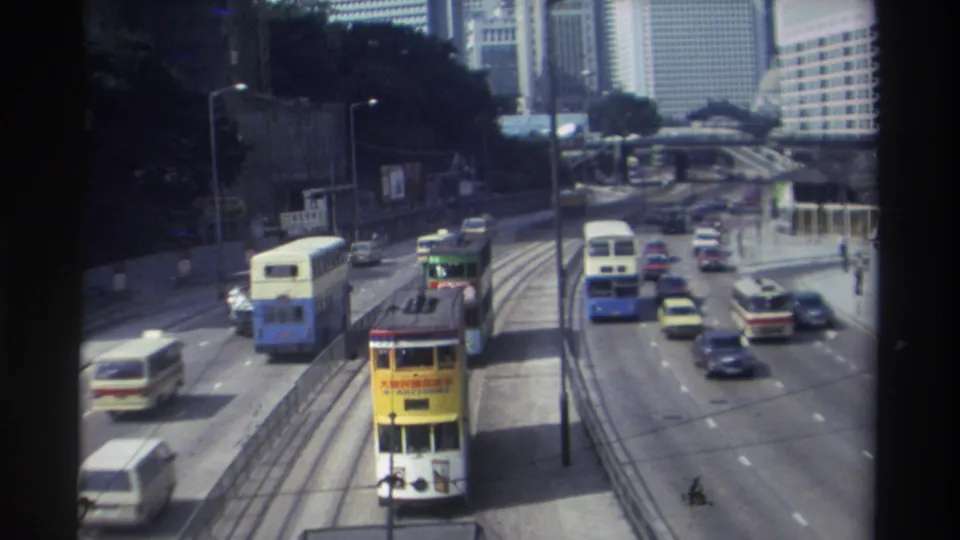 1982 Hong Kong traffic scene with a yellow double-decker tram, buses, cars, lane dividers, and tall buildings beyond the road.