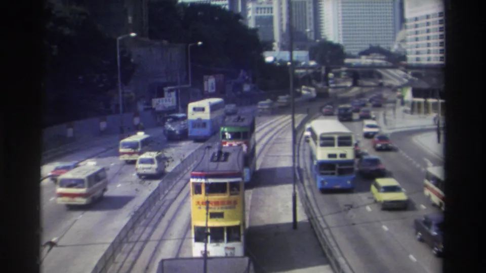 1982 Hong Kong roadway with a double-decker tram, buses, sedans, a central barrier, lamp posts, and high-rise buildings in the distance.