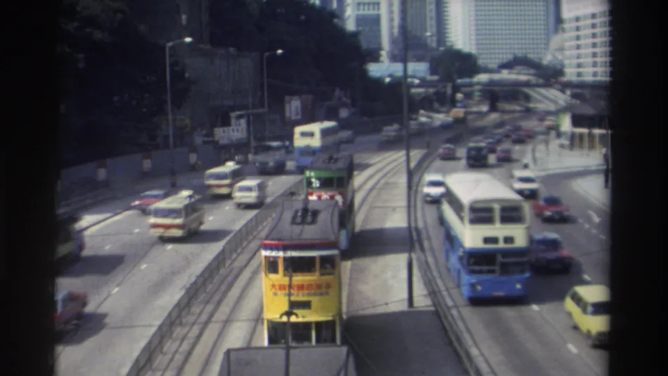 1982 Hong Kong street scene with a yellow tram in the foreground, buses and cars moving beside tracks, and tall buildings in the distance.