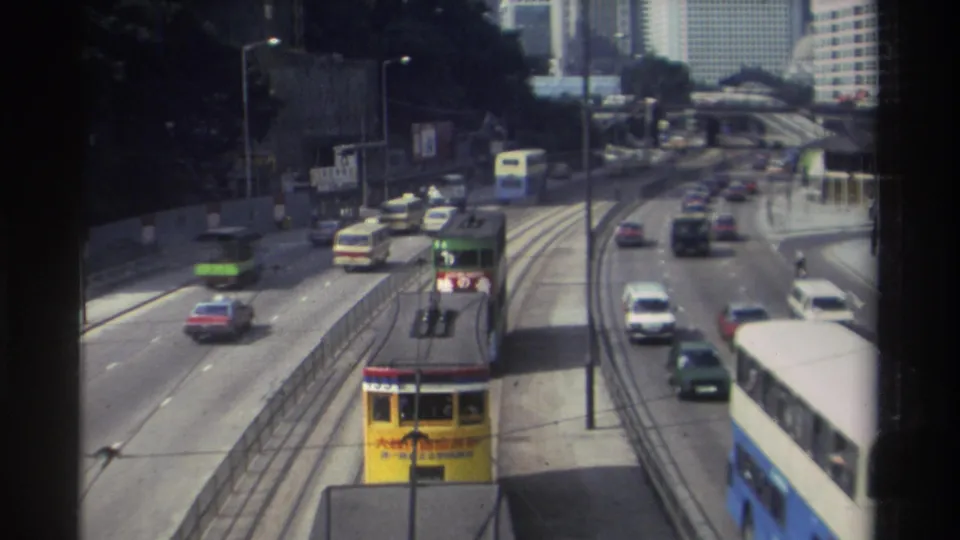 1982 Hong Kong traffic view with a yellow tram on tracks, nearby cars, buses, road signs, and high-rise buildings beyond the roadway.