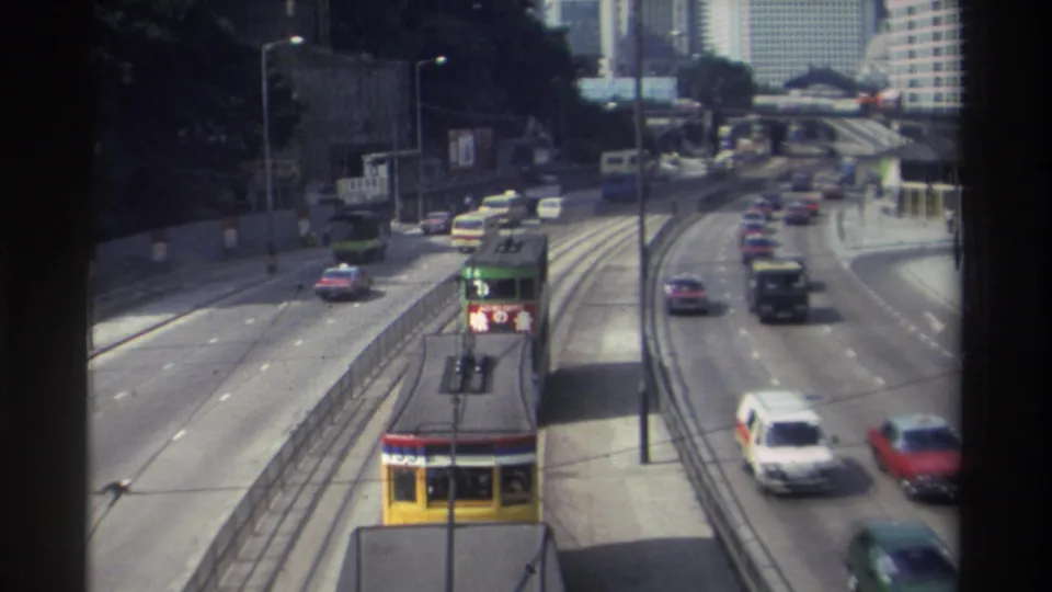 Hong Kong 1982 roadway with a yellow tram, multiple cars and vans, tram rails, road signs, and a dense high-rise background.