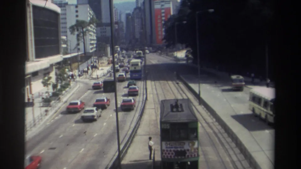 Elevated 1982 Hong Kong view with a green double-decker tram marked 105, traffic, pedestrians, a white bus, and high-rise buildings.