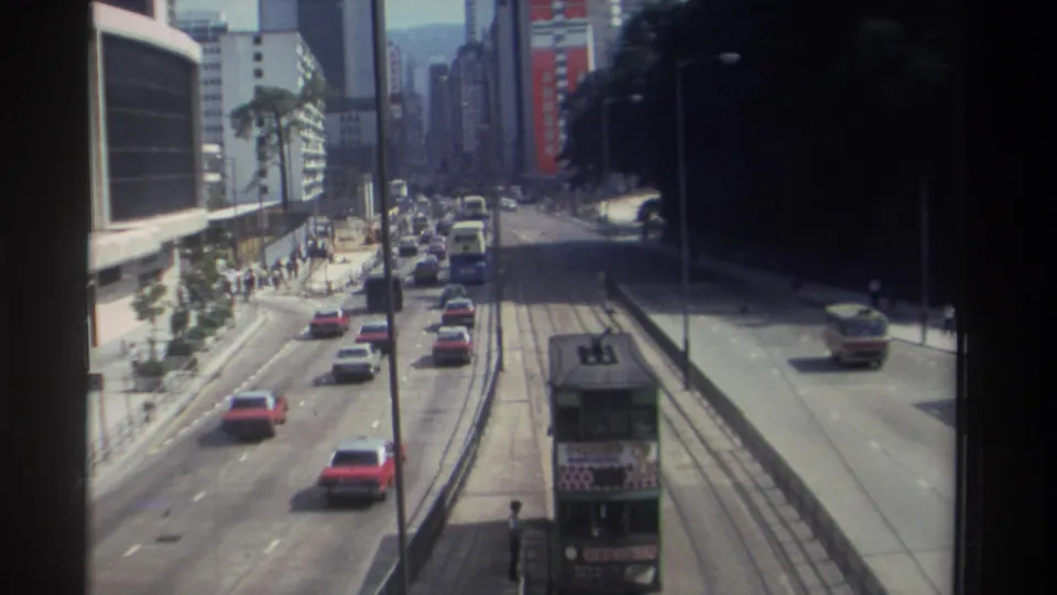 1982 Hong Kong traffic scene with a green double-decker tram on tracks, cars in nearby lanes, pedestrians on sidewalks, and dense buildings.