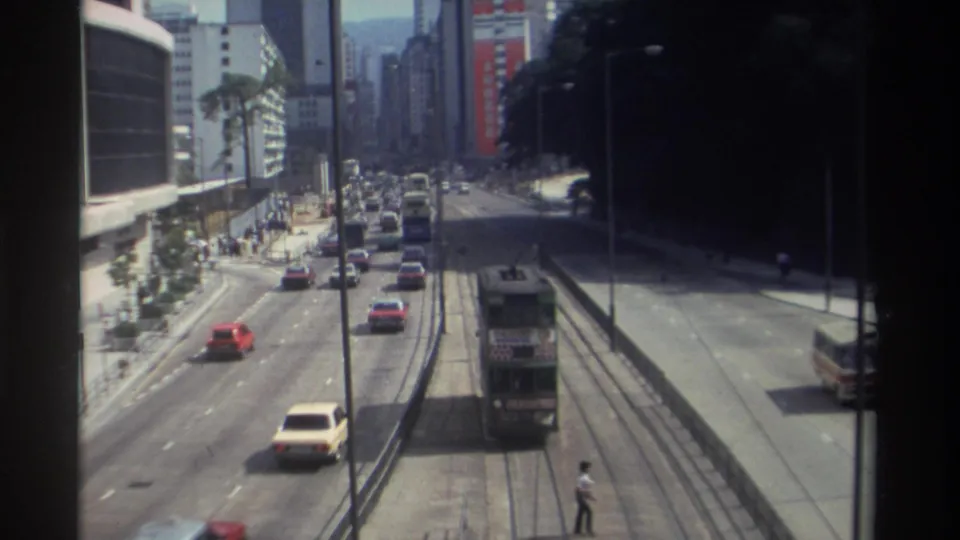 High-angle 1982 Hong Kong street view with embedded tram tracks, a double-decker tram, classic cars, and pedestrians along the sidewalks.