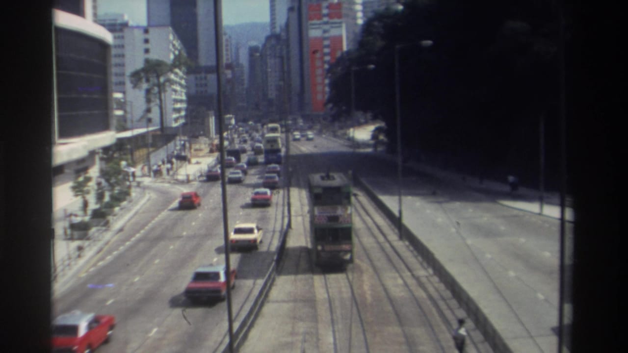 Urban road with a tram on tracks, cars in traffic, buildings, and shaded sidewalk areas.