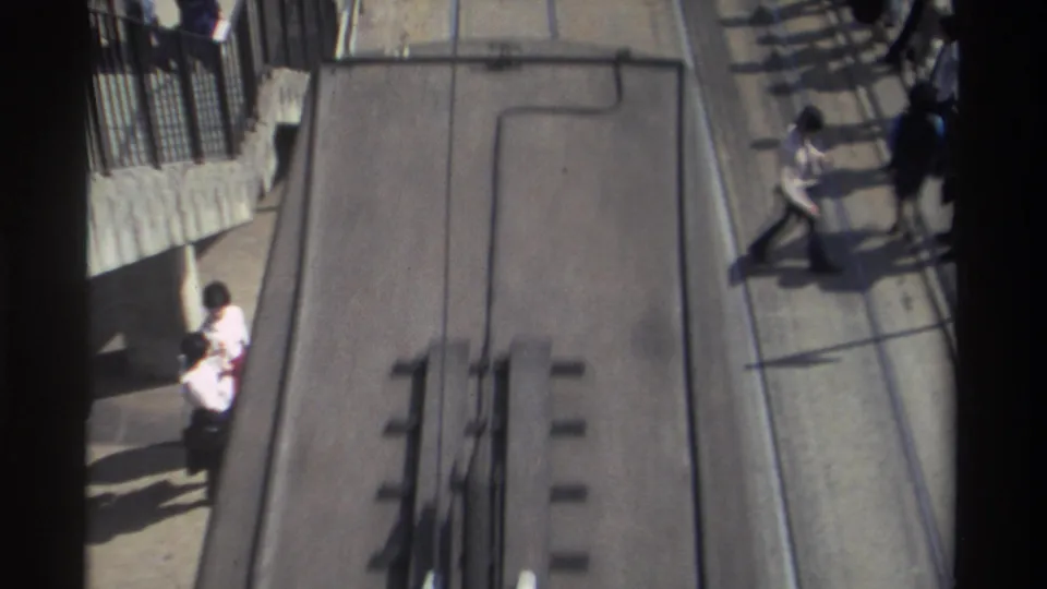 Closer elevated view of a Hong Kong tramway area, with rails, sidewalk railings, pedestrians, and strong shadows across the street surface.