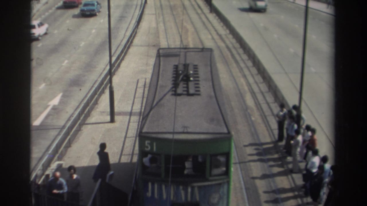 Green double-decker tram number 51 approaches passengers waiting on a 1982 Hong Kong tram platform beside active road traffic.