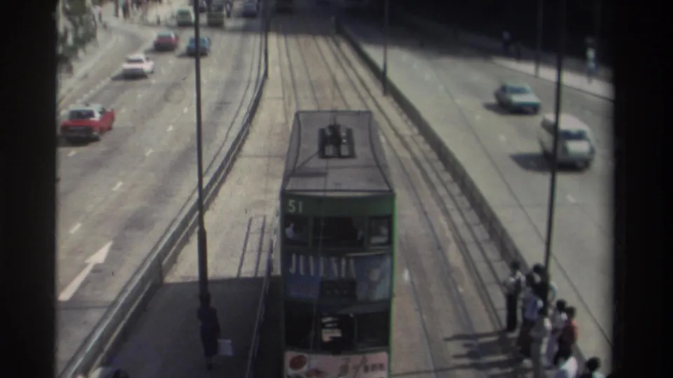 A green Hong Kong double-decker tram passes below the camera, with waiting passengers, road traffic, and overhead framing visible.