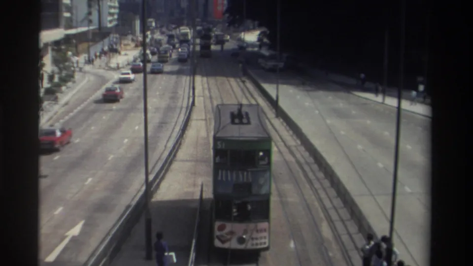 Rear high-angle view of a green double-decker tram traveling away on Hong Kong tracks amid cars and roadside buildings.