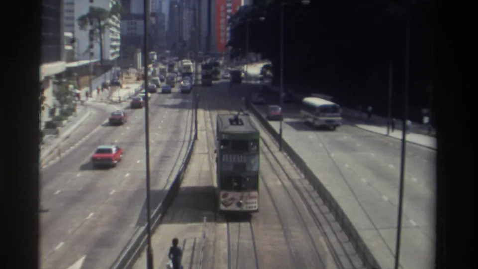 1982 Hong Kong traffic scene showing a green double-decker tram on rails, with cars, a bus, sidewalks, and tall buildings around it.