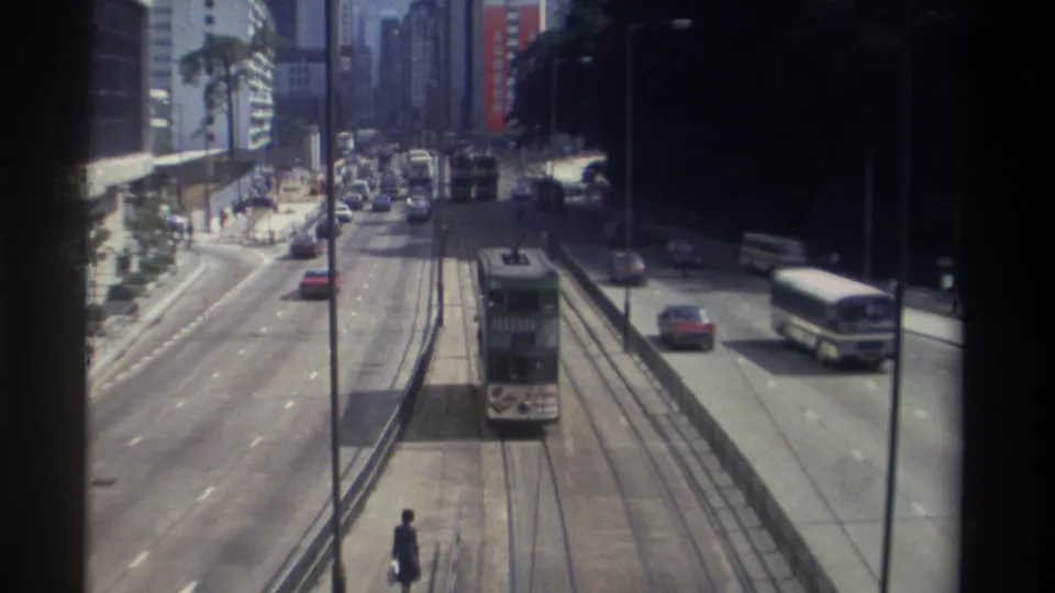 Elevated 1982 Hong Kong view with a double-decker tram on central rails, a roadside bus, cars, pedestrians, and high-rise frontage.