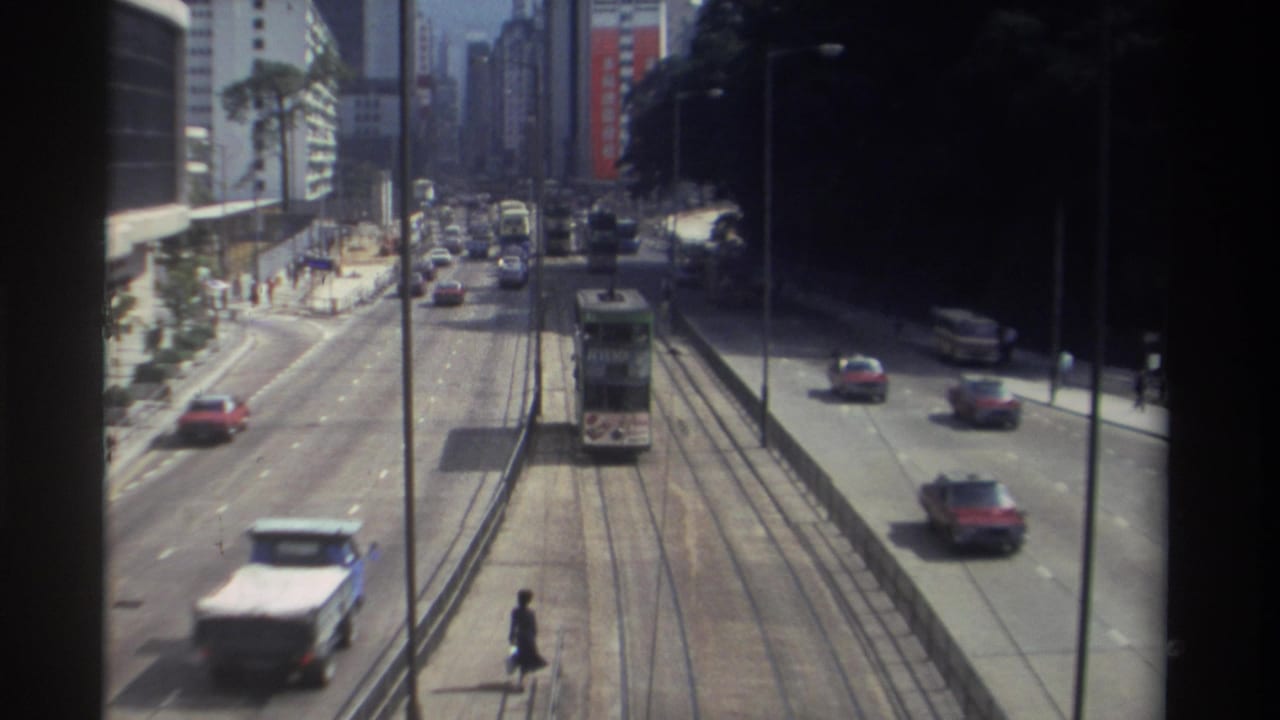 1982 Hong Kong elevated street view with a tram on central tracks, moving cars, pedestrians, and dense high-rise buildings.