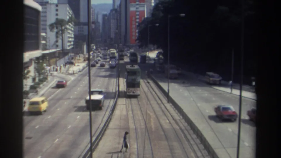 Elevated 1982 Hong Kong street scene with a tram centered on tracks, cars in several lanes, and pedestrians near the roadway.
