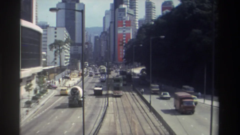 1982 Hong Kong downtown traffic from above, showing tram tracks, mixed vehicles, tall buildings, and a shaded roadside edge.