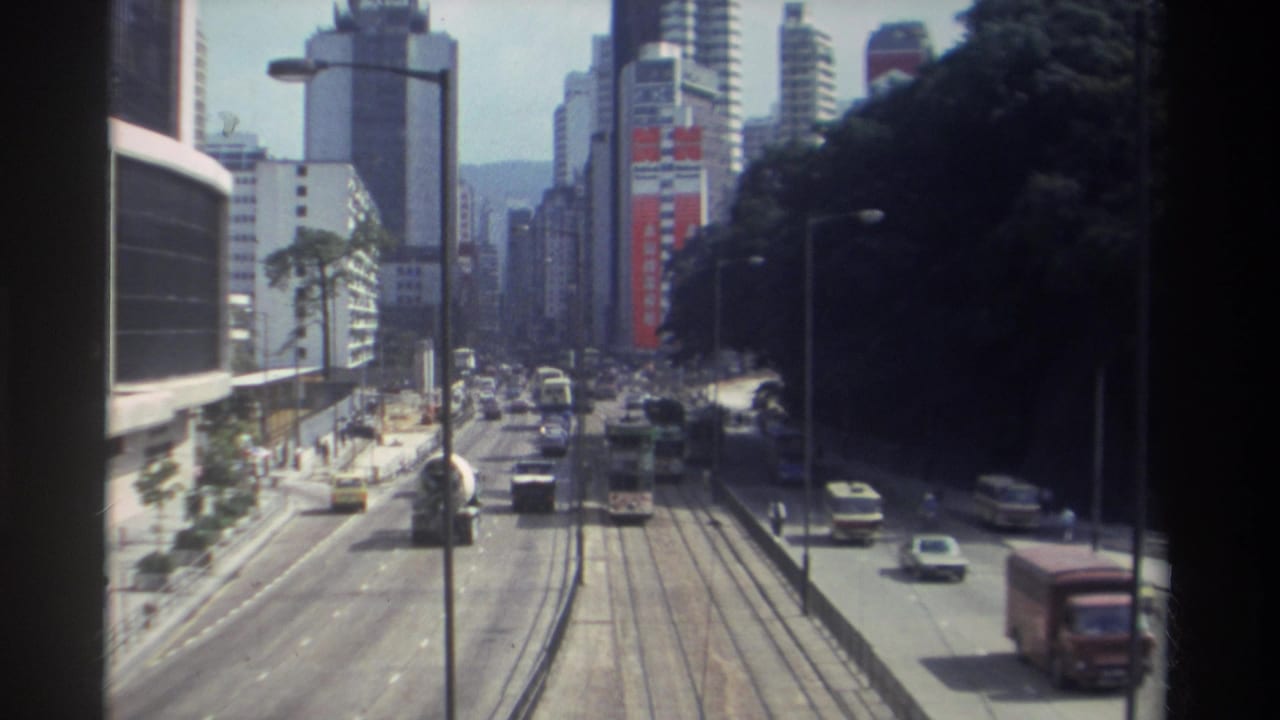 1982 Hong Kong street seen from above, with tram rails, moving traffic, buses, tall buildings, and a dark wooded hillside at right.